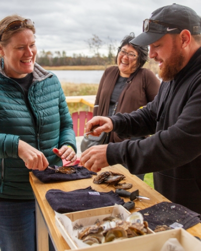 Two women learning to shuck oysters at the outdoor Oyster Deck in Cascumpec